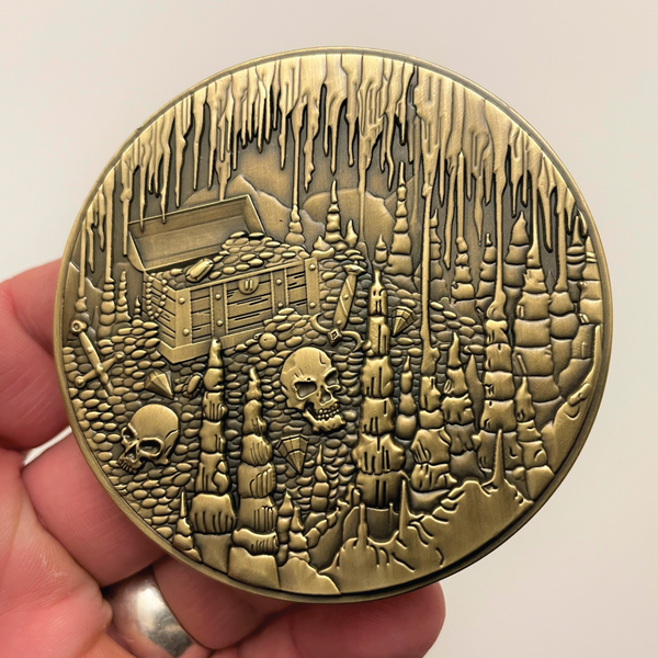 Load image into Gallery viewer, Gold metal coin in hand showing treasure chests, skulls, and caverns
