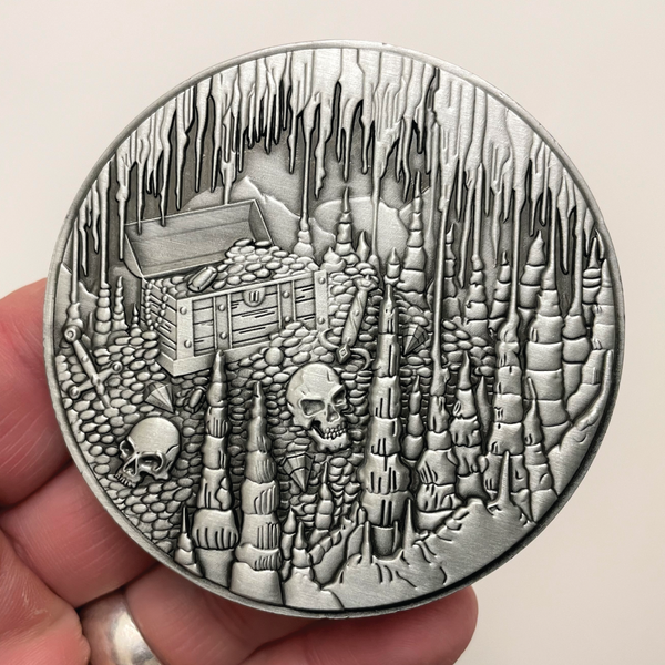 Load image into Gallery viewer, Silver metal coin with treasure chest, skulls, and cavern held in hand
