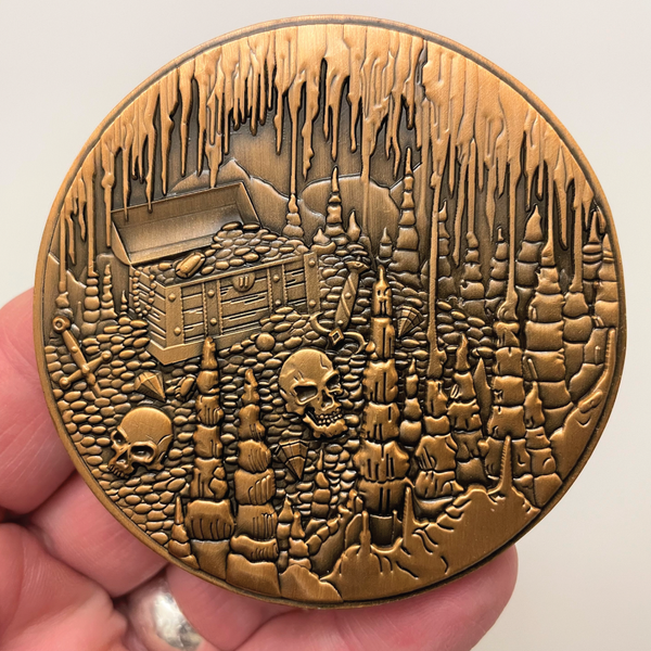 Load image into Gallery viewer, Copper metal coin in hand showing treasure chest, skulls, and cavern
