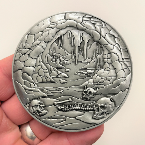 Load image into Gallery viewer, Silver metal coin in hand showing bones and cave entrance
